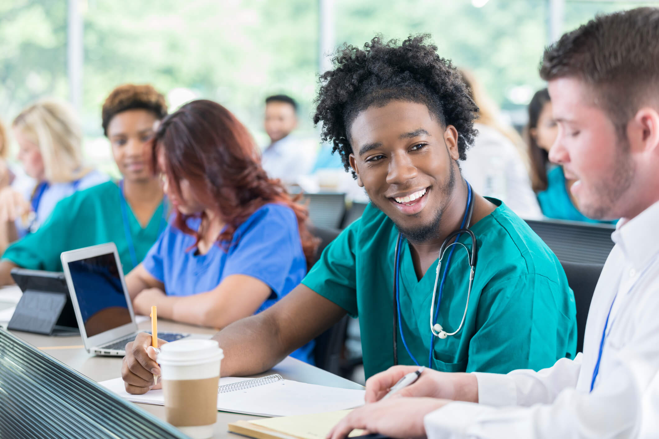 A young African American nursing school student is discussing class material with his instructor. The student is wearing teal green scrubs, and is taking notes and smiling as he listens to his instructor explain. The instructor is kneeling next to him and showing him something on a notepad. Other students are studying in the background.