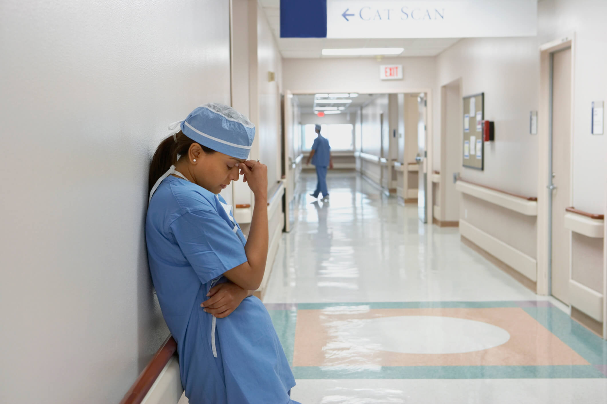 A stressed female nurse leans against a wall in a hospital hallway. Her right hand is pressed against her forehead temple, and her eyes are closed.